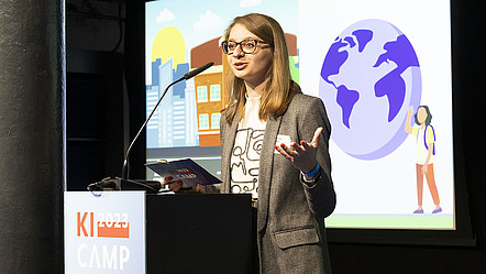 Die KI-Newcomerin Viktoriya Olari steht vor einem Podium mit der Aufschrift "KI-Camp 2023". Im Hintergrund läuft eine Präsentation zu ihrer Forschung.