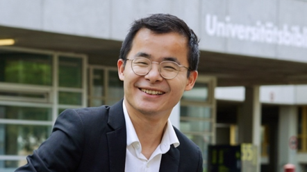 Bo Xiong trägt ein weißes Hemd und ein Sakko. Er blickt lächelnd an der Kamera vorbei. Er befindet sich vor einer Universität auf einer Wiese.
