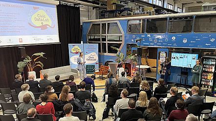 Ein Saal mit einer Bühne, auf der eine Moderatorin vor Publikum steht