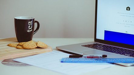 Auf einem Schreibtisch vor einem Laptop liegen ein Blatt Papier, ein Stift und ein Lineal und daneben eine Teetasse mit einigen Keksen.