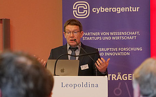 Christoph Lüth stands at a lectern at the Leopoldina and speaks into a microphone. In the background is a display of the cyber agency with a purple background.