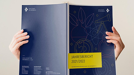 Zwei Hände halten den aktuellen Jahresbericht aufgeschlagen hoch, dahinter ist eine Person zu erkennen, die ihn liest