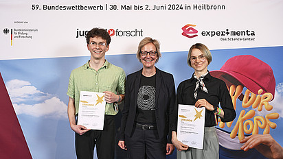 GI-Präsidentin Christine Regitz mit Alois Bachmann und Elora Marx vor der Pressewand des Wettbewerbs. Die beiden halten Urkunden in der Hand.