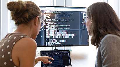 Zwei Frauen vor einem Laptop und einem Bildschirm, auf dem Code zu sehen ist