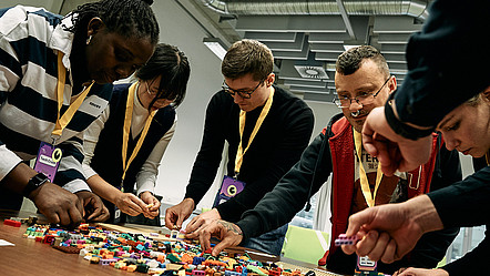 Eine diverse Gruppe junger Menschen bei einem Workshop, sie bauen Prototypen mit bunten Legosteinen.