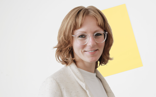 Nina lächelt in die Kamera. Sie trägt ein weißes Oberteil und einen hellen beigen Blazer.