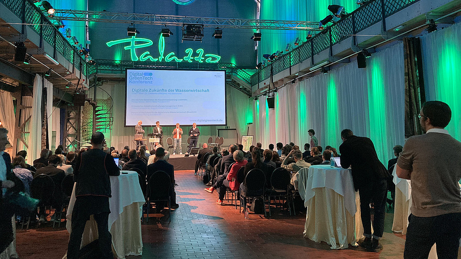Blick in eine Halle, auf deren Bühne mehrere Personen stehen. Über ihnen eine Präsentation zum Thema Digital GreenTech.