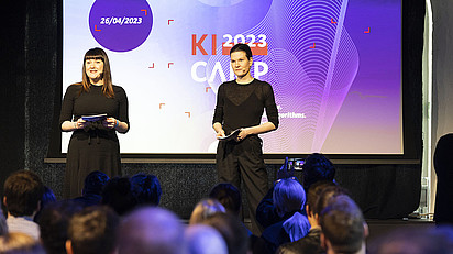 Die Projektleitung des KI-Camps Maike Klein und Julia Meisner stehen auf einer Bühne und eröffnen die Convention KI-Camp 2023. Im Hintergrund ist eine Leinwand mit dem KI-Camp Logo in den Farbtönen lila, rot und blau zu sehen.
