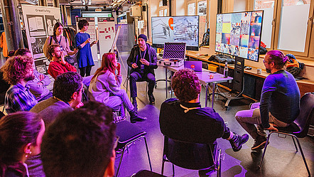 Auf dem KI-Camp 2019 sitzen Teilnehmende des Workshops in einem Halbkreis und hören dem Speaker zu. Der Speaker veranschaulicht etwas mithilfe eines Methodenkoffers und mehreren Displays.